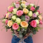 Görseldeki ürün; pembe, lila, şeftali ve krem tonlarındaki rengarenk güllerin, okaliptüs dalları ve zarif yeşil dolgu çiçekleriyle harmanlandığı, kristal ayaklı cam vazoda sunulan tam bir "bahar festivali" aranjmanıdır.