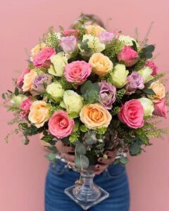 Görseldeki ürün; pembe, lila, şeftali ve krem tonlarındaki rengarenk güllerin, okaliptüs dalları ve zarif yeşil dolgu çiçekleriyle harmanlandığı, kristal ayaklı cam vazoda sunulan tam bir "bahar festivali" aranjmanıdır.