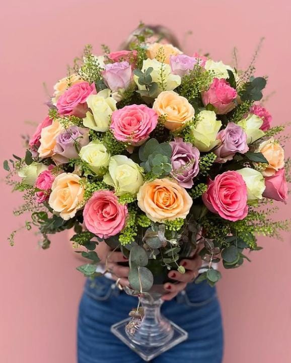 Görseldeki ürün; pembe, lila, şeftali ve krem tonlarındaki rengarenk güllerin, okaliptüs dalları ve zarif yeşil dolgu çiçekleriyle harmanlandığı, kristal ayaklı cam vazoda sunulan tam bir "bahar festivali" aranjmanıdır.