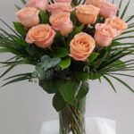 Bu görseldeki aranjman, somon/yavruağzı tonlarında ithal güller, okaliptüs dalları ve egzotik palmiye yapraklarıyla hazırlanmış, cam vazoda sunulan oldukça asil bir tasarımdır.